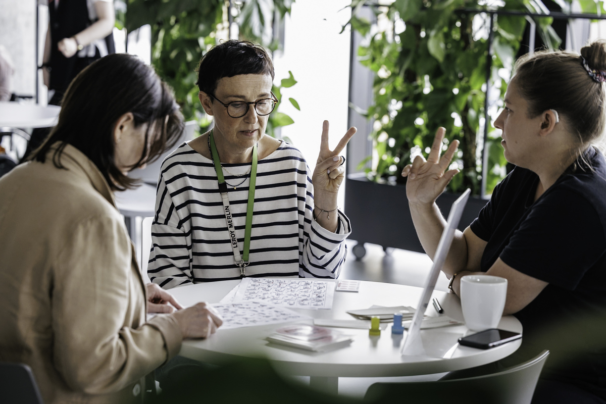 Three women sit at a table and talk using Polish sign language. On the table an educational board and a cup. In the background are green plants.